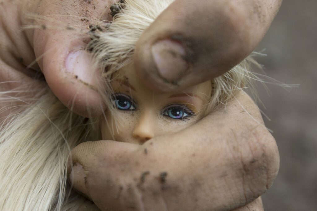 La cabeza de una muñeca rubia de ojos azules apretada por una mano de hombre sucia.