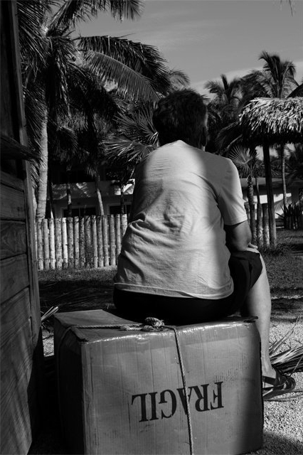Mujer anciana sentada sobre una caja de cartón en que se lee "Frágil"