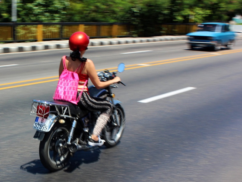 Mujer conduce una motocicleta llevando un casco rojo y una jaba rosada a la espalda.