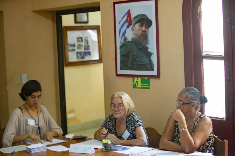 Tres mujeres trabajando en las elecciones en Cuba, con cuadro de Fidel detrás.
