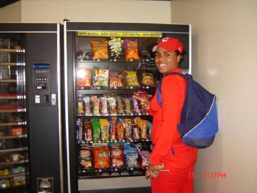 Fotografía de Raisa de Feria, jugadora de béisbol femenino con su uniforme frente a un dispensador de confituras.